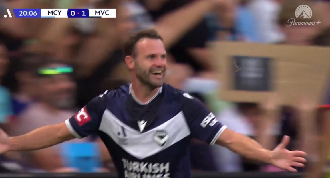 A soccer player in a dark blue and white jersey with "TURKISH AIRLINES" on it celebrates, smiling widely with arms outstretched. The score "MCY 0-1 MVC" is visible.