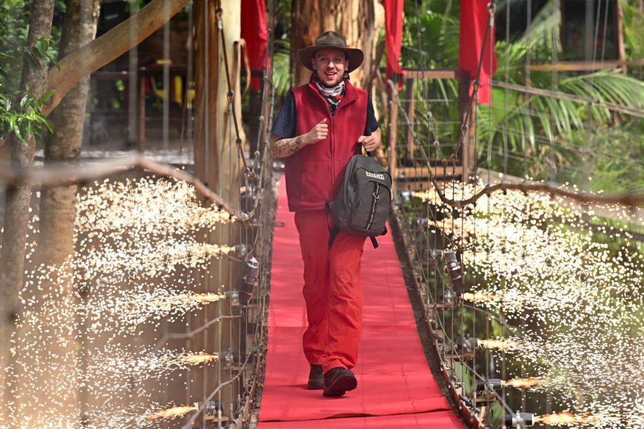 A man in a red vest and pants, carrying a backpack, walks across a red bridge with sparks flying from the sides.