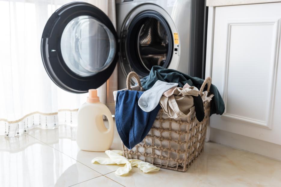 An open washing machine door, a basket of laundry, a bottle of detergent, and a pair of white socks on the floor.