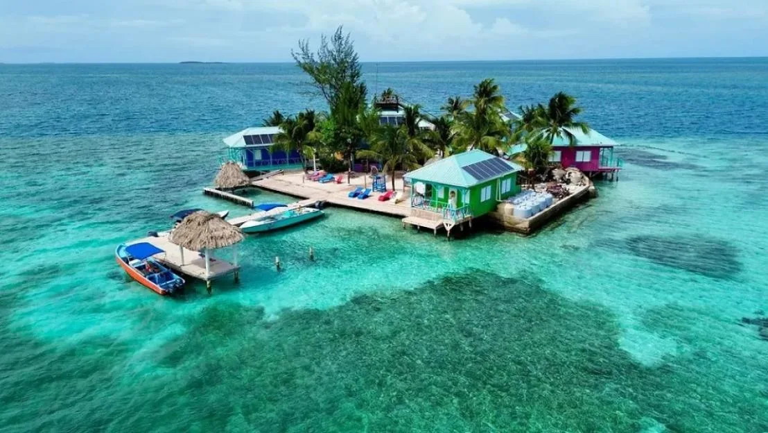 Aerial view of King Lewey's Island Resort in Belize, surrounded by turquoise water and coral reefs, with colorful buildings, a dock, and boats.