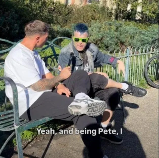 Two men sit on a park bench in front of the Brighton Museum and Art Gallery, with one man interviewing the other who wears blue-dyed hair and neon sunglasses.
