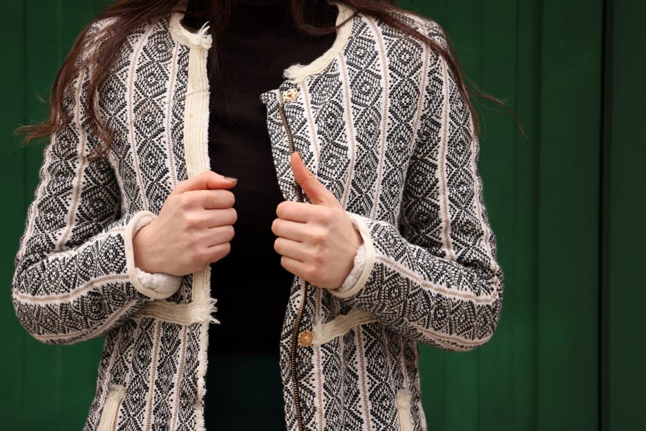 A smiling woman with long dark hair, wearing a denim jacket and ripped jeans, walks on a city sidewalk holding a coffee cup and a black bag.