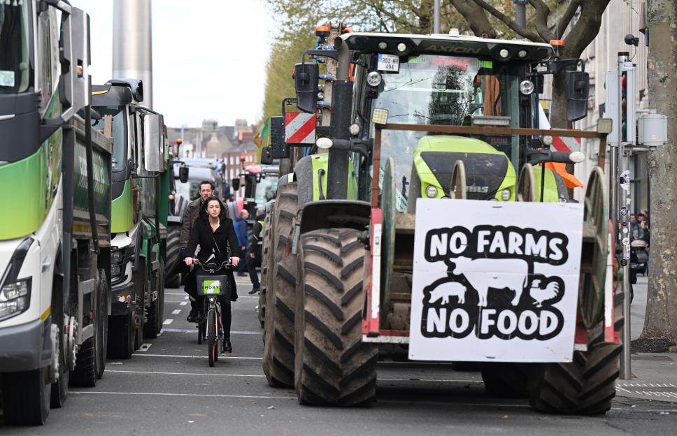 A woman on a bicycle rides past parked tractors and trucks, with a protest sign reading "No Farms No Food" visible on the back of a tractor.