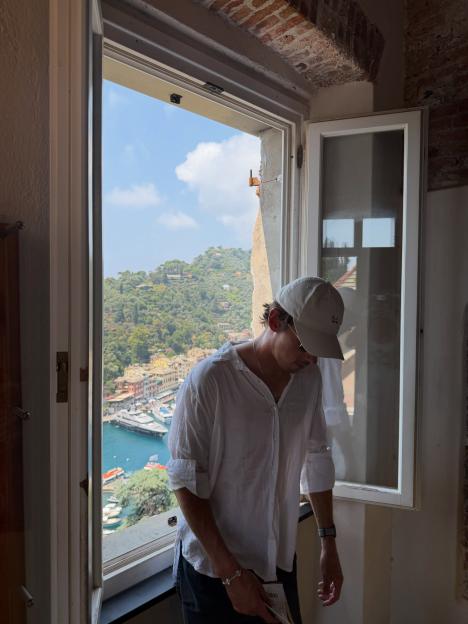 Man in white shirt and hat looking down, with a view of a harbor town and green hills behind him.