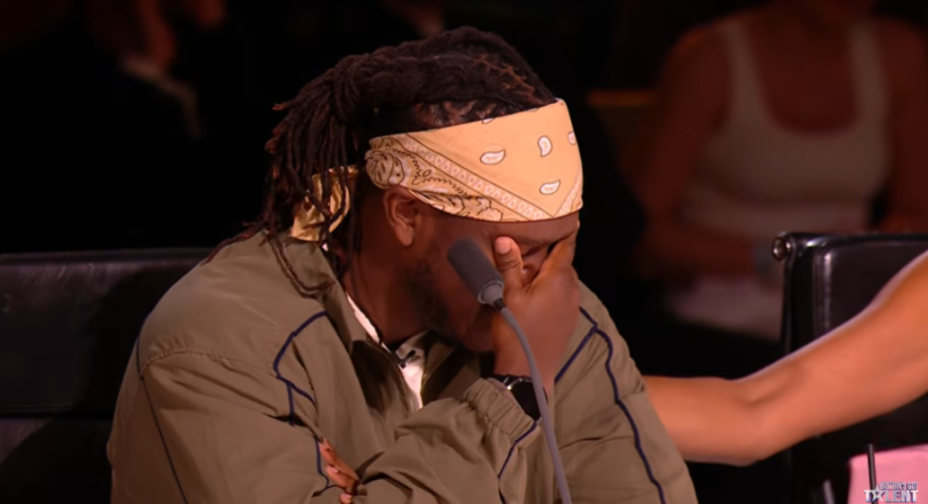 A man with dreadlocks and a bandana covering his eyes, appearing emotional, with a microphone in front of him.