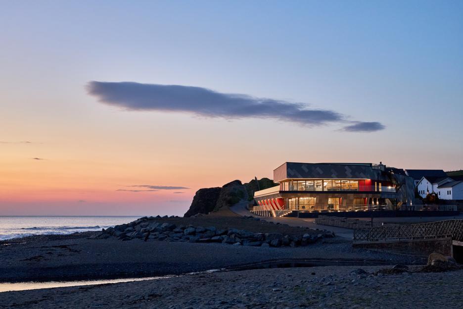 A building with illuminated windows and red accents on a cliff by the ocean at sunset.
