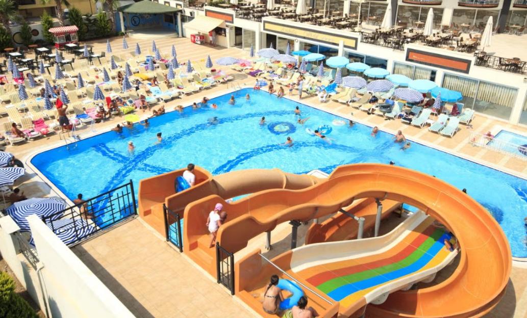 Overhead view of a bustling resort swimming pool with a large waterslide, surrounded by lounge chairs, umbrellas, and buildings.