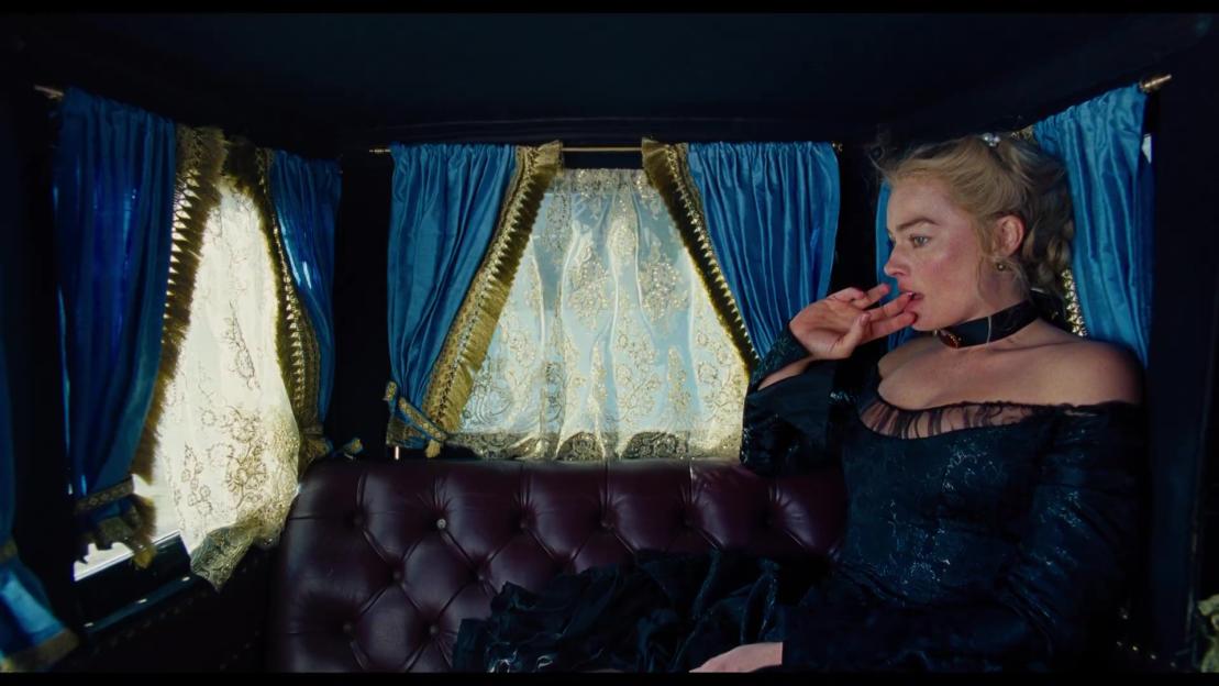 Margot Robbie in a black off-the-shoulder gown, sitting in a carriage with blue and gold curtains.