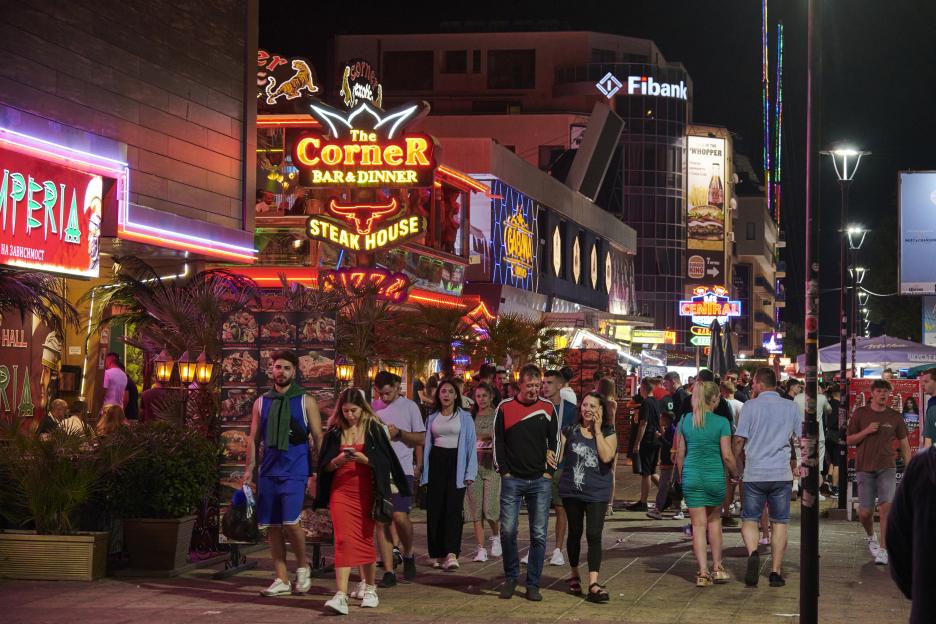People walking on a busy street lined with brightly lit restaurants and shops at night.