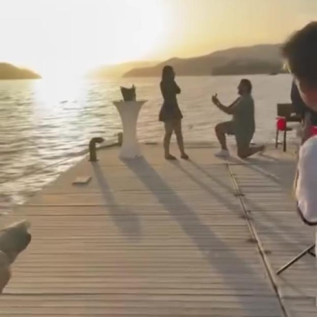 A man kneels on a dock to propose to a woman, with the sun setting over the water in the background.