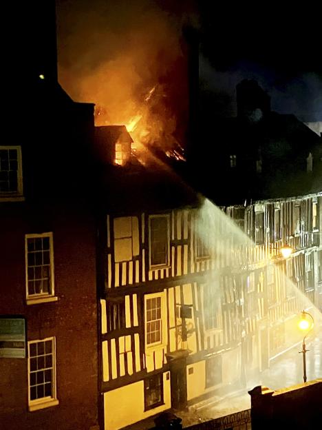 Firefighters spray water on a fire blazing from the roof of a historic timber-framed building at night.