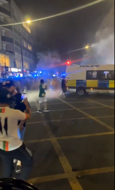 Crowd dispersing as police deploy tear gas from a police van on a street at night.