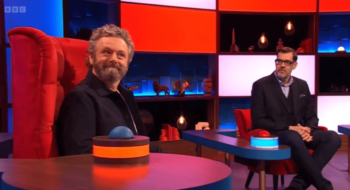 Michael Sheen sitting in a large red chair with a buzzer on the desk in front of him, next to Richard Osman on a TV set.
