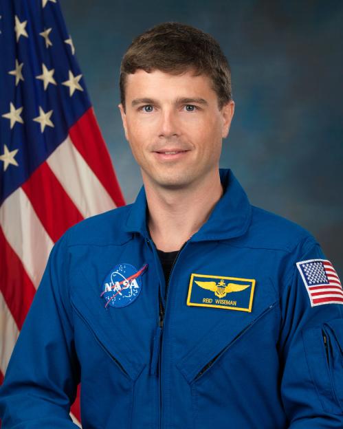 Astronaut Reid Wiseman in his blue flight suit in front of an American flag.