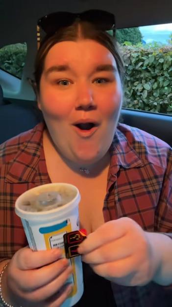 Becki Jones in a car, surprised, holding a drink and a small item.