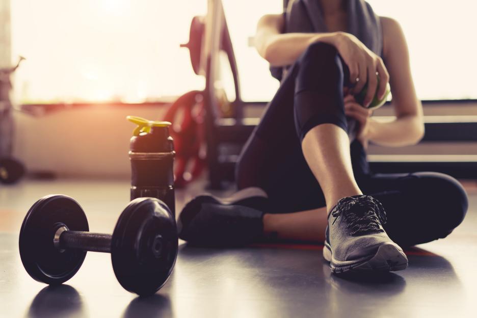 A woman in workout clothes sits on the gym floor, holding an apple, next to a dumbbell and water bottle.