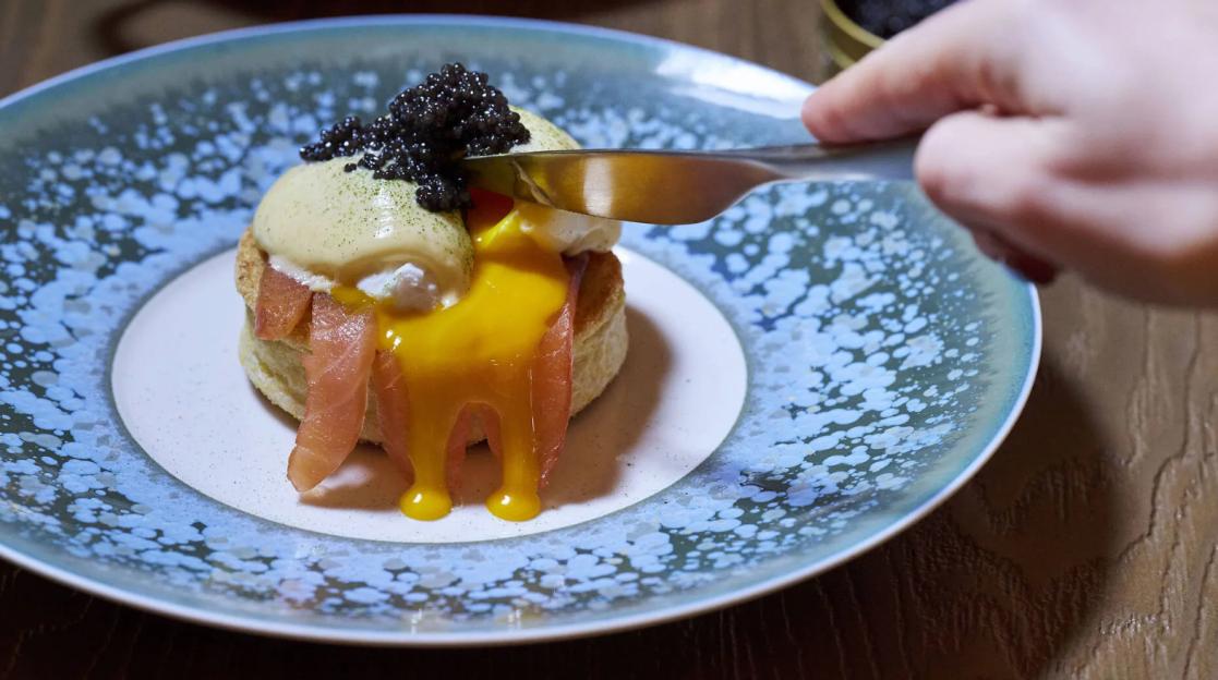 A chef's knife cuts into a breakfast dish with caviar, hollandaise sauce, and smoked salmon on a biscuit, as egg yolk oozes out.