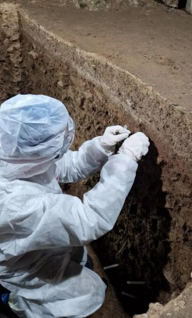 A person in a white protective suit and gloves taking a sample from the side of an excavated trench.