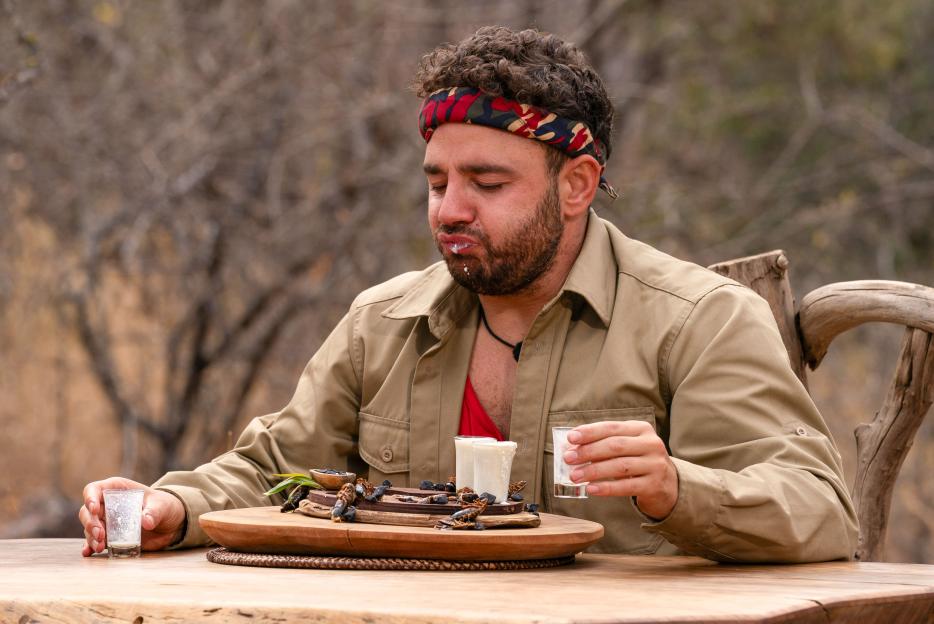 A man, wearing a camouflage bandana and khaki shirt, eats food from a wooden platter with a white substance on his face during a bushtucker trial from "I'm A Celebrity... Get Me Out Of Here! South Africa".