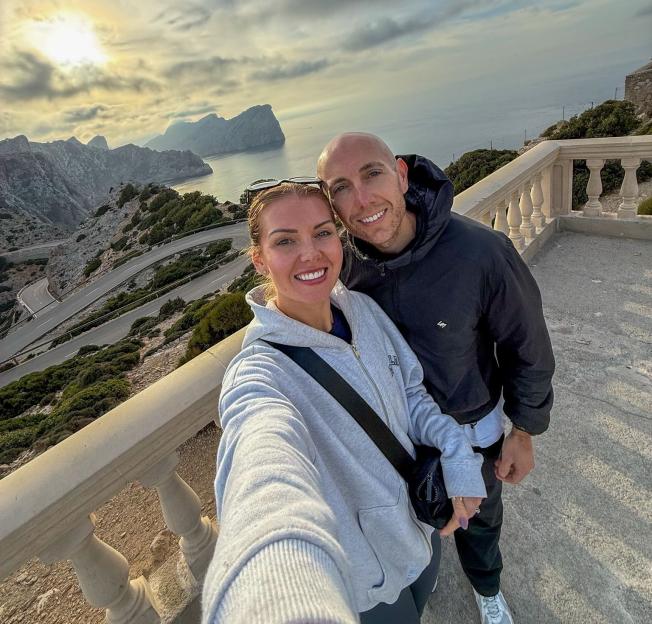 Jordan and Ray, a married couple, posing for a selfie in Majorca with mountains, a winding road, and the sea in the background.