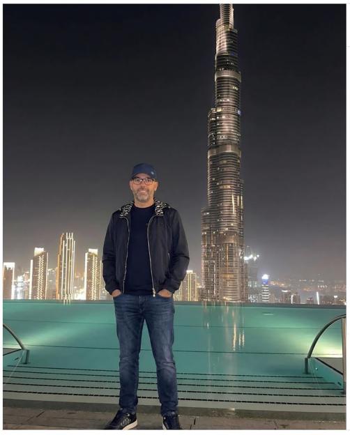 Man in a jacket, jeans, and cap standing by a pool with the Burj Khalifa in the background at night.