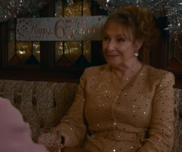 Woman in a sparkly gold jacket, with a "Happy 65th Birthday" banner behind her.