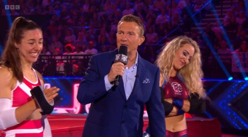 Bradley Walsh holding a microphone between two female Gladiators.