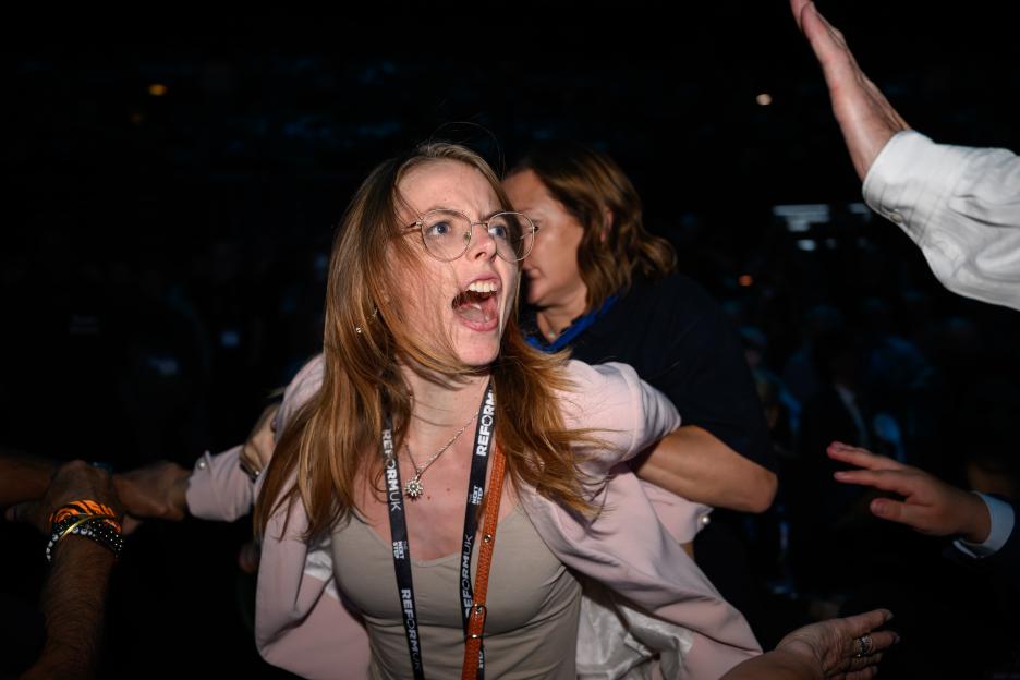 A protester with glasses and open mouth is being removed from a political event.