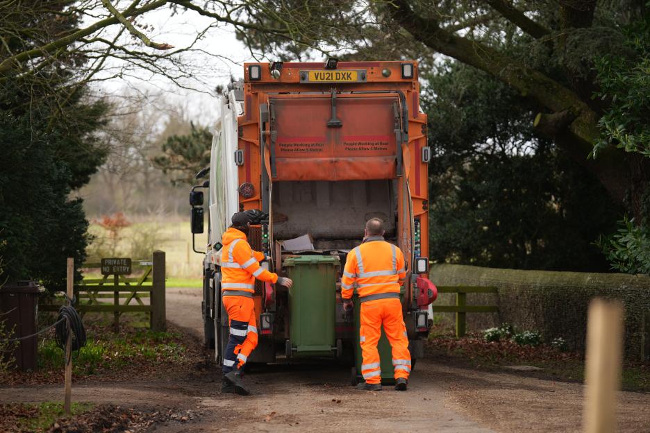 Two waste collectors in orange uniforms emptying a green bin into the back of a garbage truck.