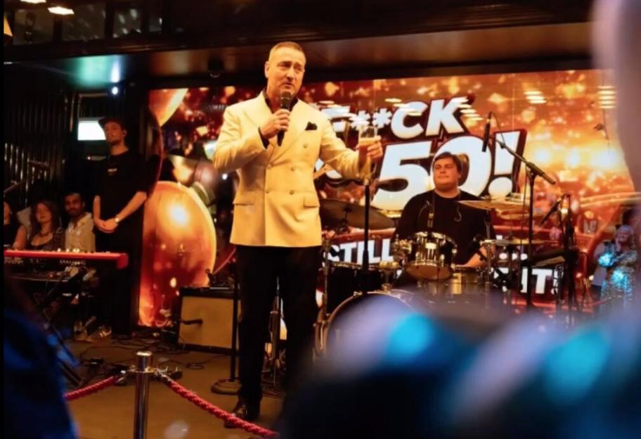 Will Mellor speaking into a microphone with a glass of champagne in hand at his 50th birthday party.