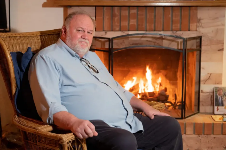 An older man with a white beard and glasses hanging from his shirt collar sits in a wicker armchair in front of a lit fireplace.