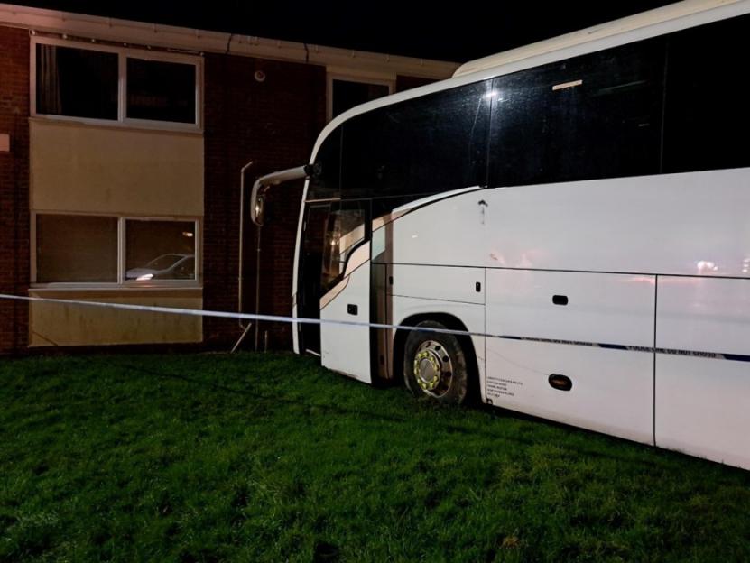 A large white coach bus with a damaged side is seen on a grassy area next to a brick building, cordoned off by "POLICE LINE DO NOT CROSS" tape.