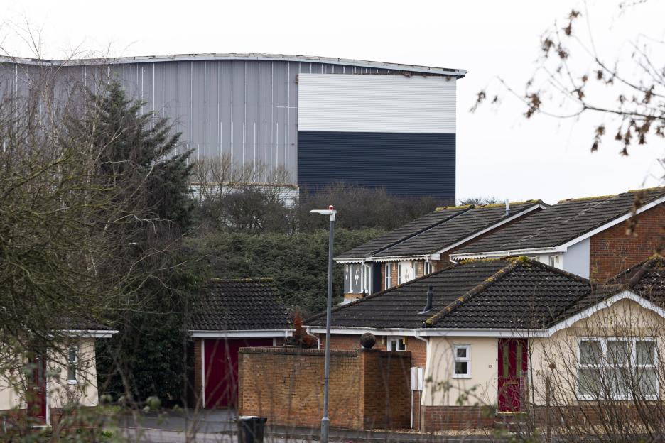 A large warehouse with grey, white, and navy siding looms over residential houses and trees.