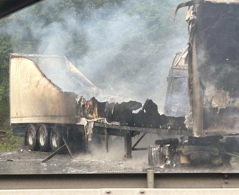 A burnt semi-truck trailer is smoldering on the side of a road, with dark smoke rising against a backdrop of green trees.