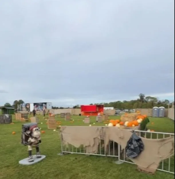 A sparse pumpkin patch featuring a character statue, hay bales, and fencing in a grassy field under an overcast sky.