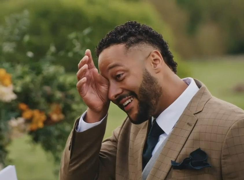 A man in a checkered suit wipes a tear from his eye while smiling, showing emotion.