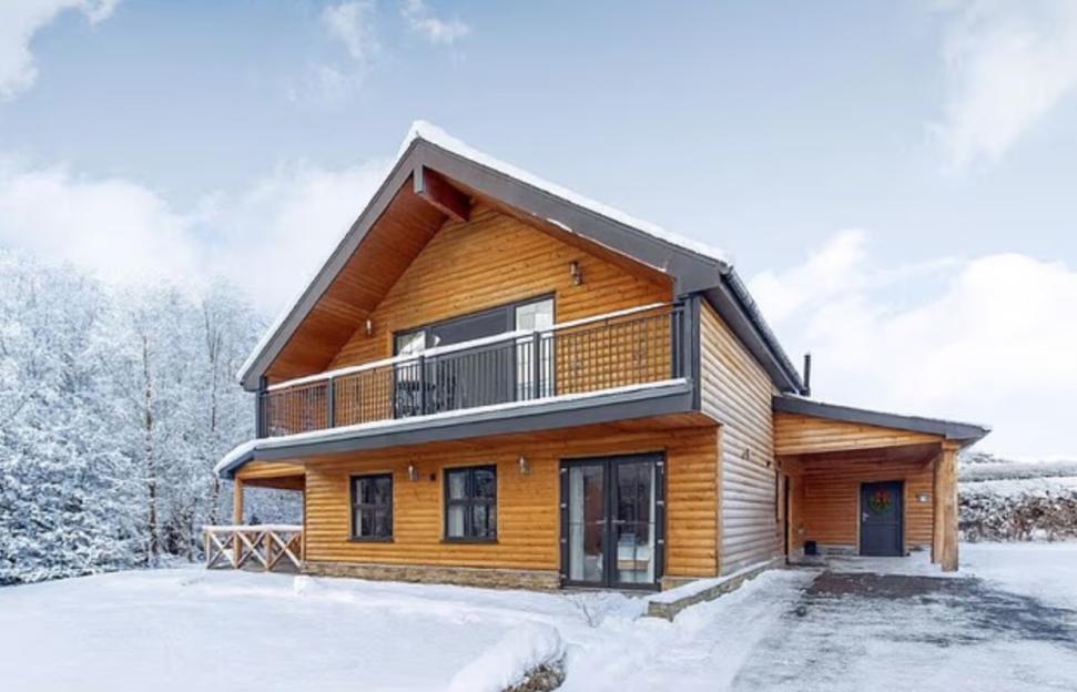 A wooden holiday lodge with a balcony and covered entryway, surrounded by snow and snow-covered trees.