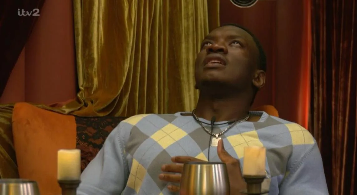 A Black man in a blue and yellow argyle sweater looks upward in disbelief, holding one hand to his chest.