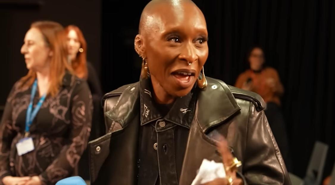 Bald person wearing a leather jacket and large earrings speaks.