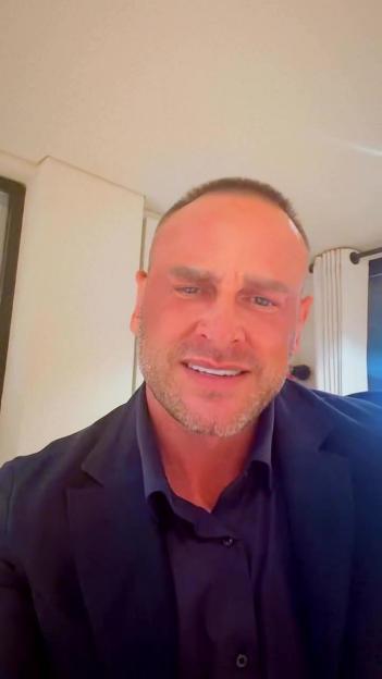 A bald man in a black shirt and suit jacket with stubble and a slight smile.