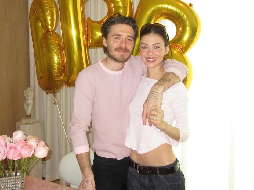 Nicola Peltz and Brooklyn Beckham pose with their arms around each other in front of golden balloons and a bouquet of pink roses.