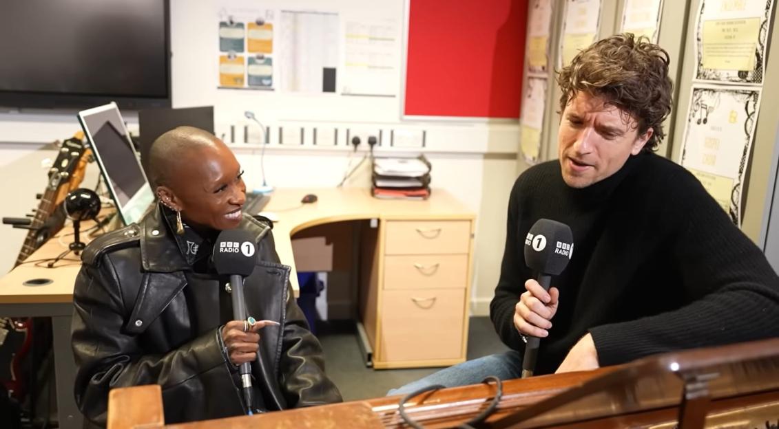 Two people, one in a leather jacket and another in a black turtleneck, holding BBC Radio 1 microphones.