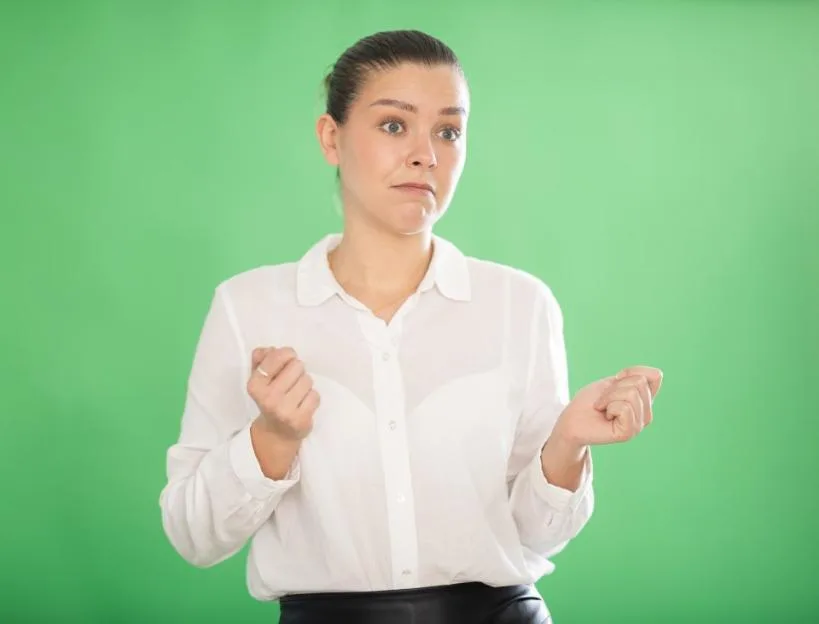 A young woman stands against a green background, gesturing thoughtfully with her hands with a confused expression on her face.
