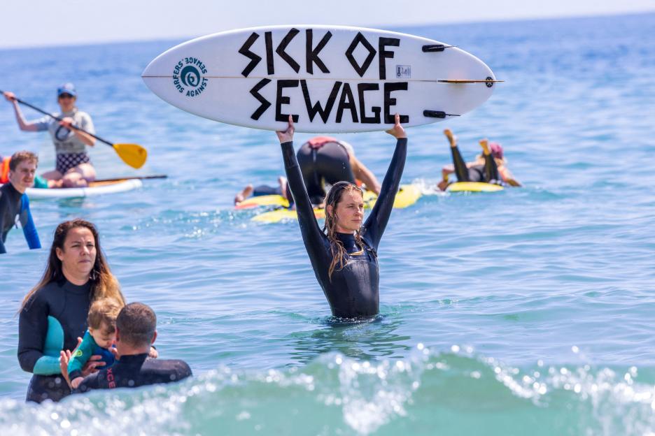 A woman holding a surfboard above her head that says "SICK OF SEWAGE" while standing in the ocean during a paddle-out protest.
