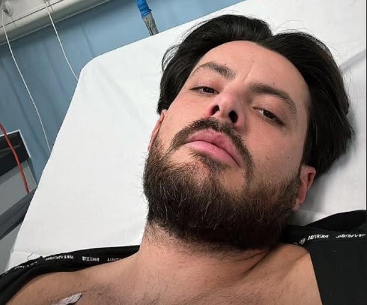 A man with dark hair and a beard lies in a hospital bed, looking at the camera.