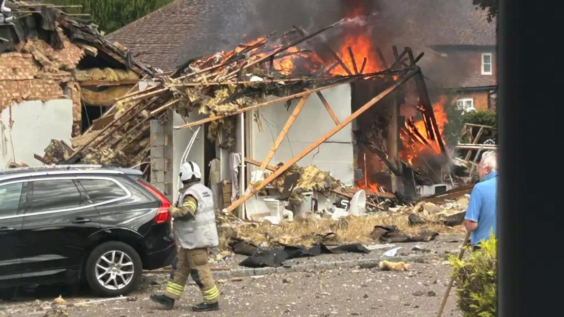A destroyed bungalow with an active fire, smoke, and debris, with a firefighter in the foreground.