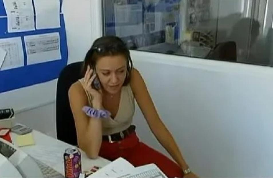 A woman with dark hair, wearing sunglasses on her head, a light-colored top, and red pants, sits at a desk while talking on a phone.