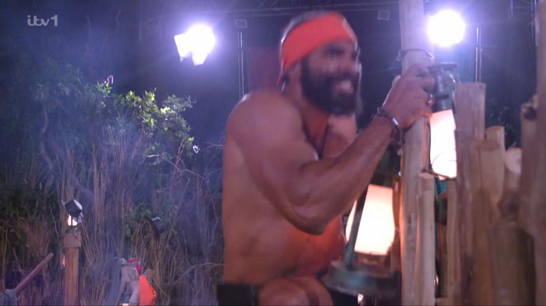 A shirtless man with a red headband looking distressed, holding onto a wooden structure in a dark, outdoor setting with bright lights.