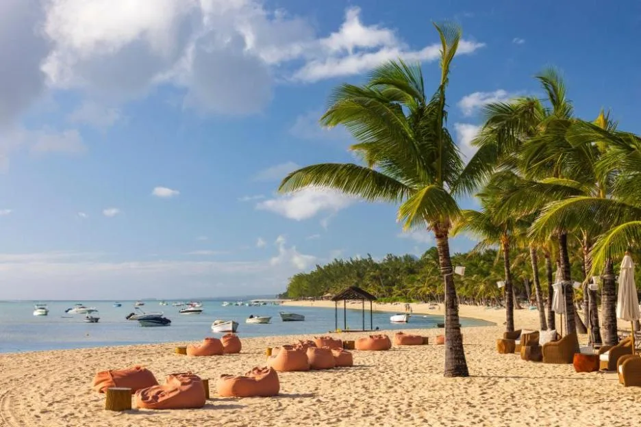 Tropical beach at Le Morne in Mauritius with palm trees, sandy shore, bean bag chairs, and boats in the ocean under a blue sky.