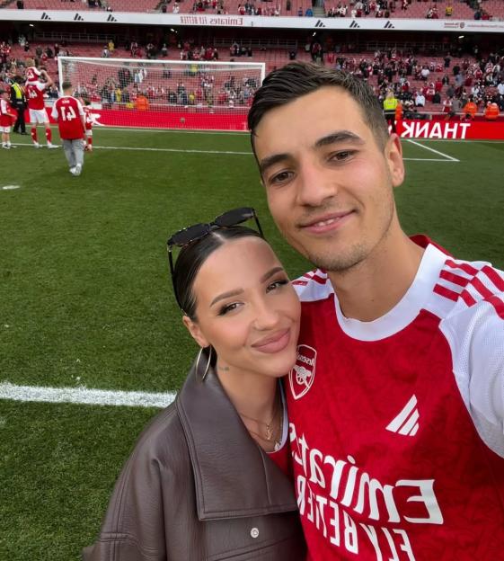 Jakub Kiwior and his wife Claudia Kowalczyk posing on a soccer field in front of a goal and stadium seating.